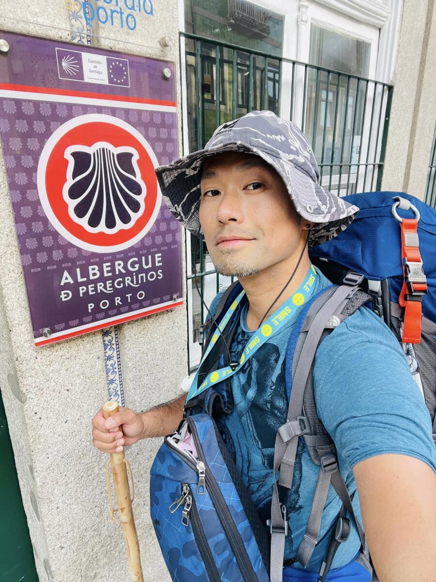 史坦力背著背包站在 Albergue de Peregrinos Porto 的紫色扇貝招牌前