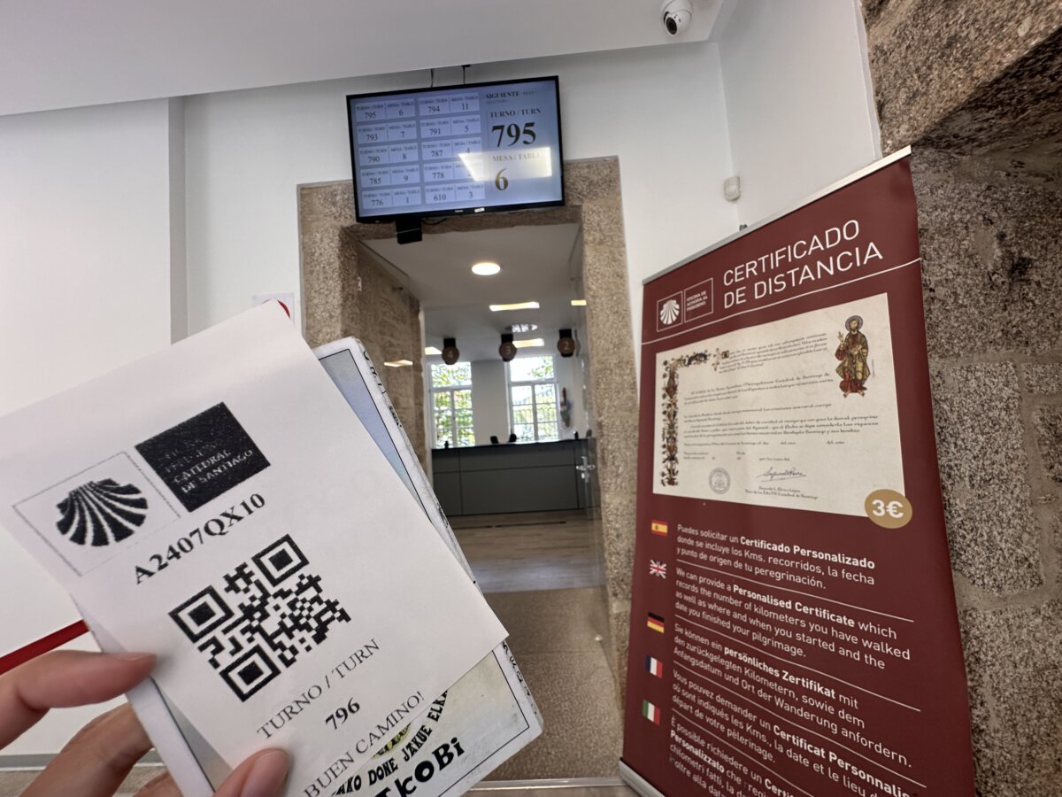 朝聖者辦公室內，手拿印有 QR code 的號碼牌，背後是公里數證書公告牌與辦公室走道