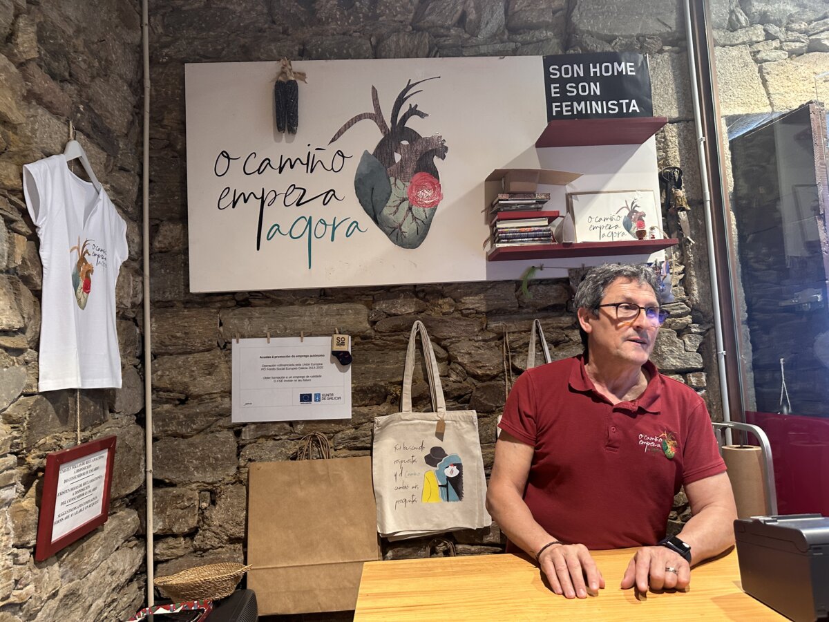 o camiño empeza ahora 紀念品店老闆坐在櫃檯，牆上掛著手繪 T 恤與帆布袋