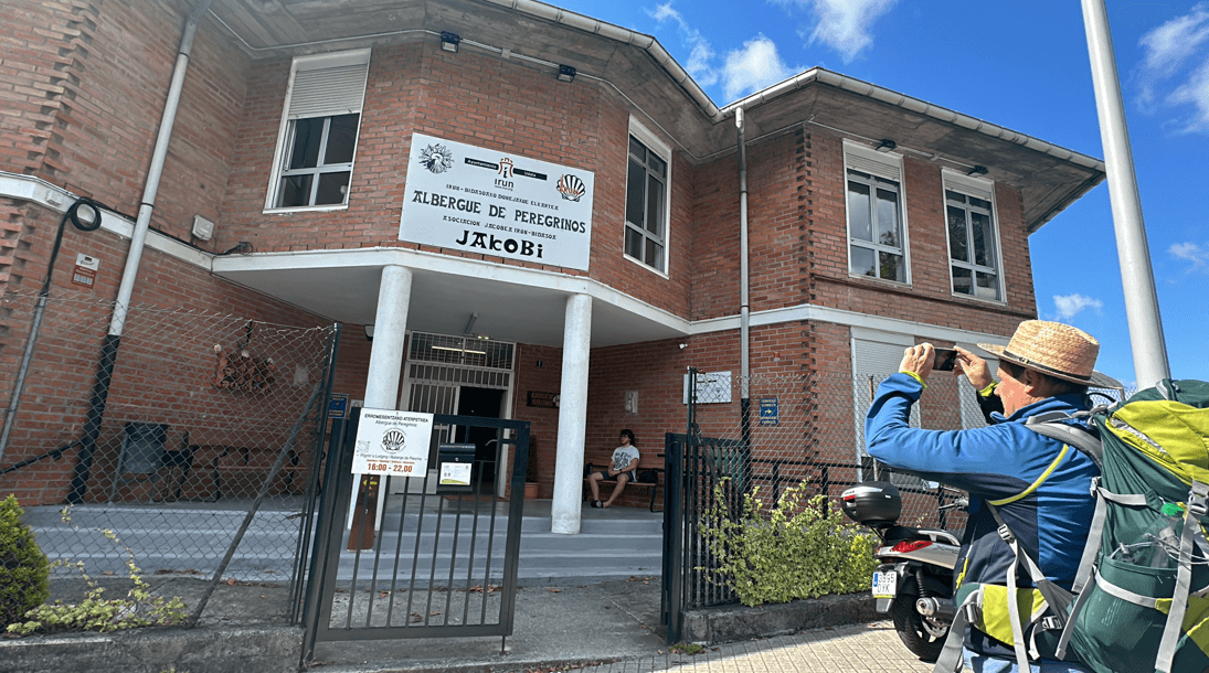 Irún 公立庇護所 Albergue de Peregrinos Jakobi 建築外觀，招牌寫著 JAKOBI，一位朝聖者背著登山包在門口拍照