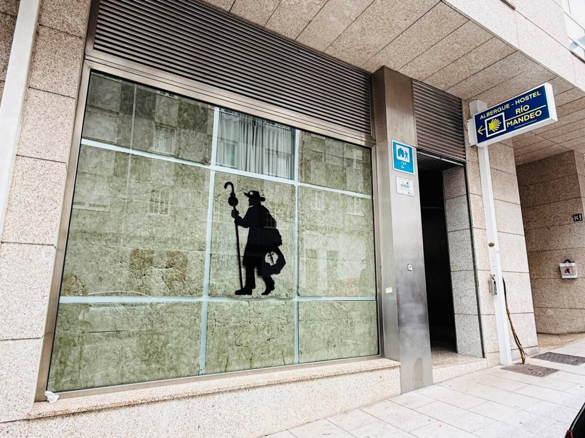 Betanzos 的 Albergue Río Mandeo 門口，牆上掛著藍色 ALBERGUE HOSTEL RÍO MANDEO 招牌和朝聖貝殼，玻璃窗上有朝聖者剪影