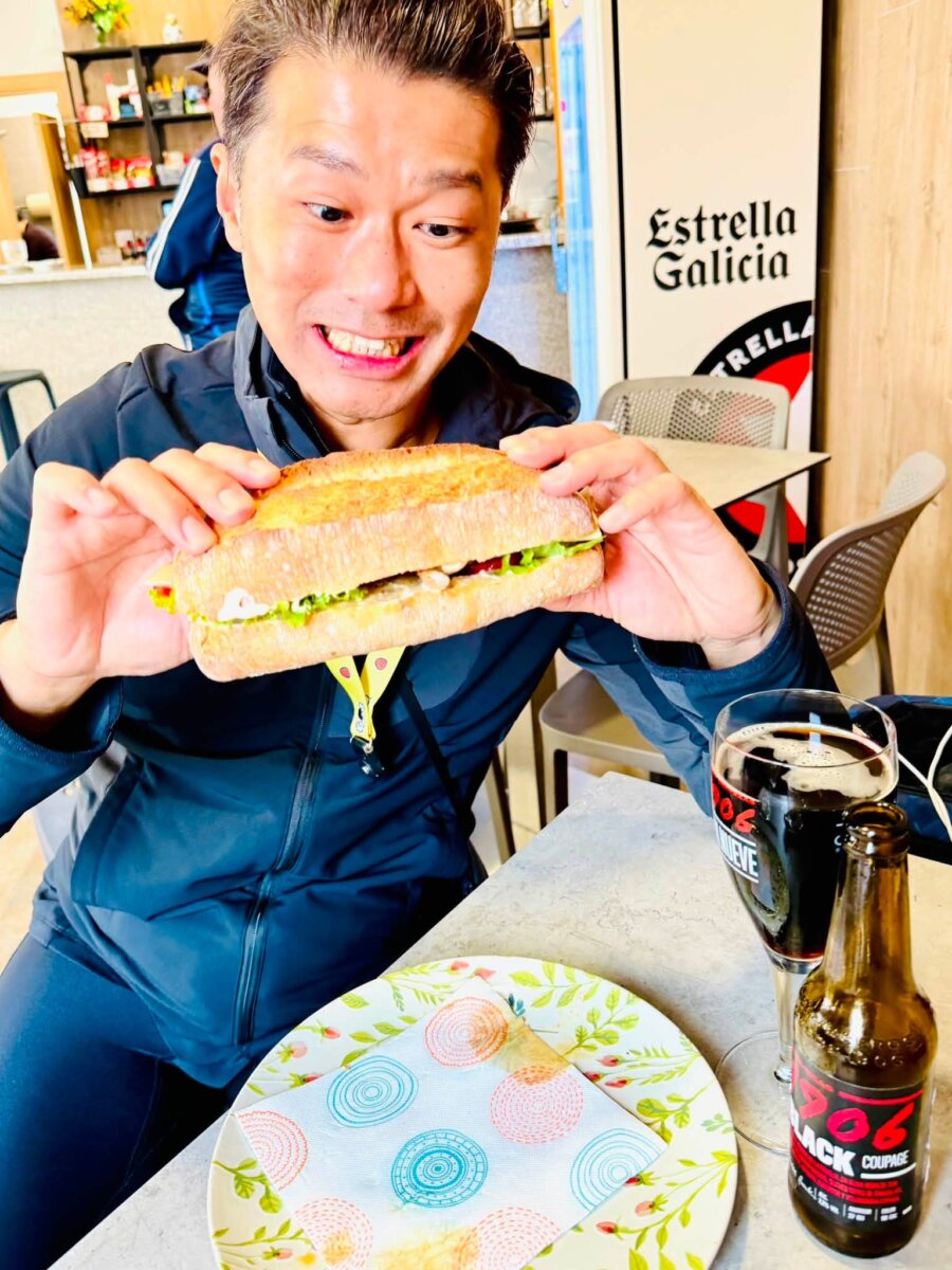 史坦力雙手捧著巨無霸三明治，歐式長麵包夾著豬排和生菜，桌上有一瓶 Estrella Galicia 啤酒